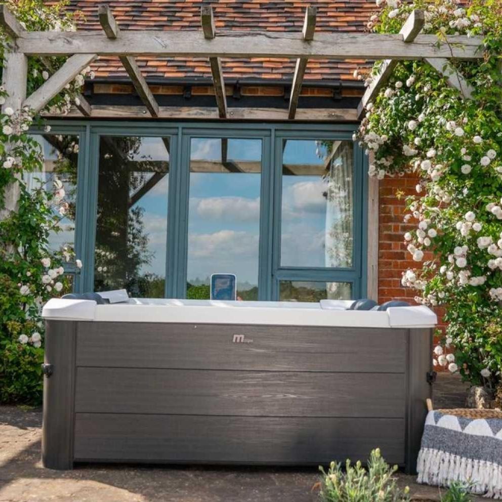 Portable grey hard frame hot tub with wood-textured paneling, set outdoors in front of glass doors surrounded by climbing. - Home Interior Shop - MSpa Oslo 4-6 Person Portable Hard Frame Hot Tub - Grey